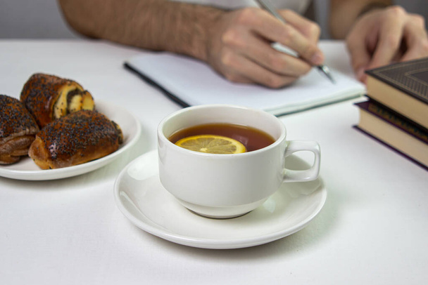 Ένα φλιτζάνι τσάι με λεμόνι σε ελαφρύ φόντο. Σνακ στη δουλειά. Ένα φλιτζάνι τσάι στη δουλειά. Ένα φλιτζάνι τσάι με γλυκά. - Φωτογραφία, εικόνα