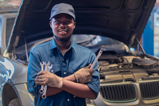 African man mechanic in uniform with crossed arms and wrenches standing at the car repair station - Foto, Imagem