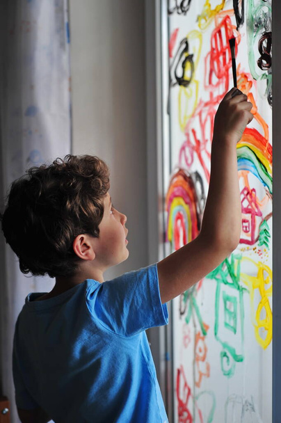 Un ragazzino serio tiene un pennello tra le mani e disegna dei disegni sulla finestra della sua stanza. Il bambino disegna un arcobaleno, una casa e un'astrazione. - Foto, immagini