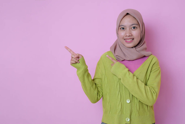 コピースペースの脇に魅力的な幸せな若いイスラム教徒のアジアのティーンエイジャーの笑顔ポイントは、広告がピンクの背景にイスラム教徒のカジュアルな服を着て示しています。プロモーションコンセプト - 写真・画像