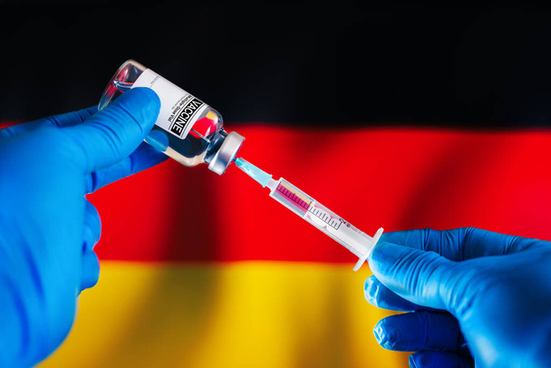 Ο γιατρός ετοιμάζει το φιαλίδιο της ένεσης του εμβολίου για το σχέδιο εμβολιασμού κατά των ασθενειών στη Γερμανία. Προετοιμασία της δόσης του εμβολίου σε σύριγγα για πρόληψη λοιμώξεων μπροστά από τη σημαία της Γερμανίας - Φωτογραφία, εικόνα