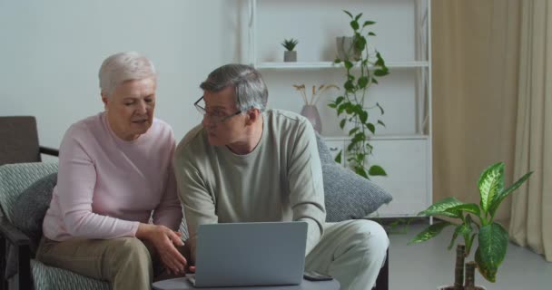 Happy old middle aged caucasian old couple using laptop technologies talking together doing online shopping choose product reading discussing internet news ordering booking tickets on website at home - Footage, Video