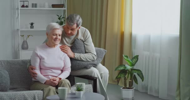 Anziani coppia caucasica si siede sul divano grigio, vecchio marito amorevole abbraccia la moglie conforta amata donna triste si sente dispiaciuto si scusa sostiene partner in situazione difficile dà aiuto psicologico - Filmati, video