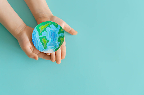 Child is holding planet in hands on teal background. Earth day holiday concept. - Fotó, kép