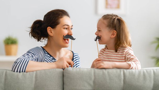 Mom and her child daughter are playing at home. Cute girl are holding paper mustache on stick. Family holiday and togetherness. - Φωτογραφία, εικόνα
