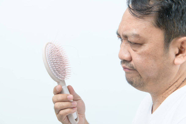 Hombre de mediana edad mirando cepillo de peine con pérdida de cabello y estresado por sus problemas de pérdida de cabello aislados en el fondo blanco, concepto de cuidado de la salud - Foto, Imagen