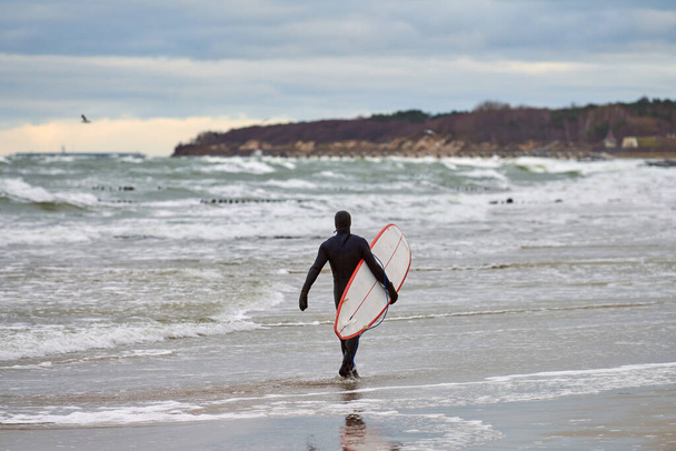 Surfeur masculin en maillot de bain noir marchant le long de la mer et tenant une planche de surf blanche dans sa main. Jour chaud, beau ciel nuageux, scène de la nature. Concept sport et activités de plein air. - Photo, image