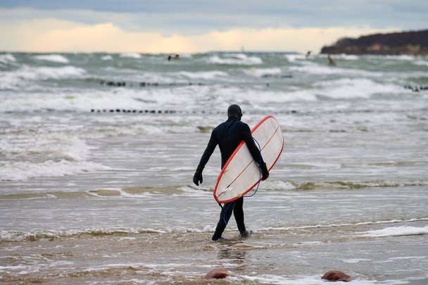 Surfista masculino en traje de baño negro caminando a lo largo del mar y sosteniendo una tabla de surf blanca en su mano. Día cálido, hermoso cielo nublado, escena de la naturaleza. Concepto de deporte y actividades al aire libre. - Foto, imagen