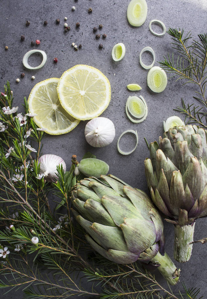 Fresh artichokes on a table. Top view photo of uncooked vegetables: artichoke flowers, lemon slices, leeks and herbs. Still life food. Balanced  diet concept.  - Photo, Image
