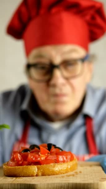 Senior volwassen chef-kok Kaukasische etniciteit in rode chef-kok hoed versiert bruschetta met basilicum tomaten in de huishoudelijke keuken. Focus op de voorgrond. Selectieve focus. Artistieke wazige achtergrond. - Video
