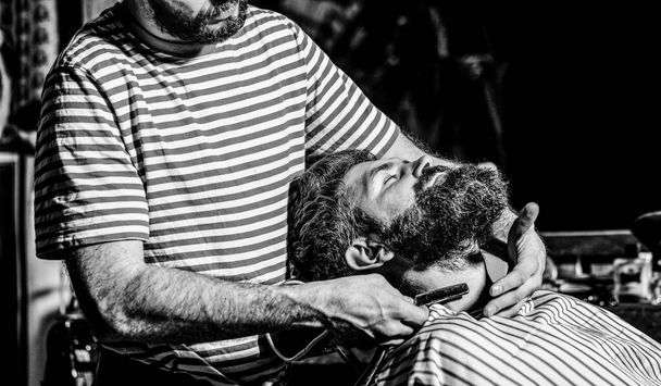 床屋の椅子に腰掛けている髭の男は、美容師の髭を危険なカミソリで剃っている。理髪店で髭の男を剃る理髪師. - 写真・画像