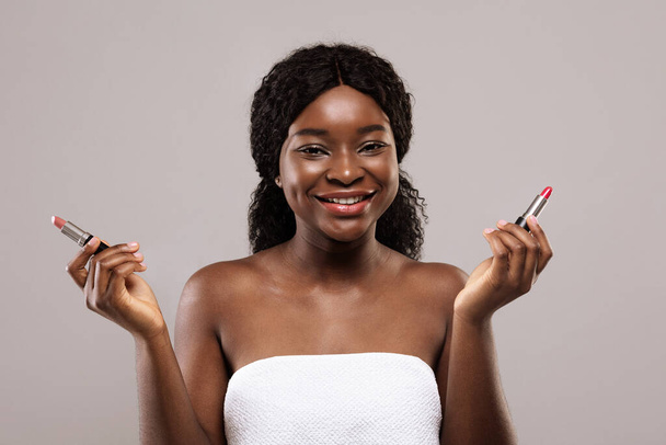 Sorridente giovane donna nera avvolta in asciugamano con due rossetti in mano - Foto, immagini