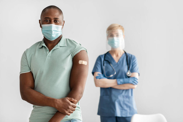 Ώριμη νοσοκόμα ή γιατρός με στολή, προστατευτική μάσκα στέκεται με χιλιετή αφροαμερικανός ασθενής - Φωτογραφία, εικόνα