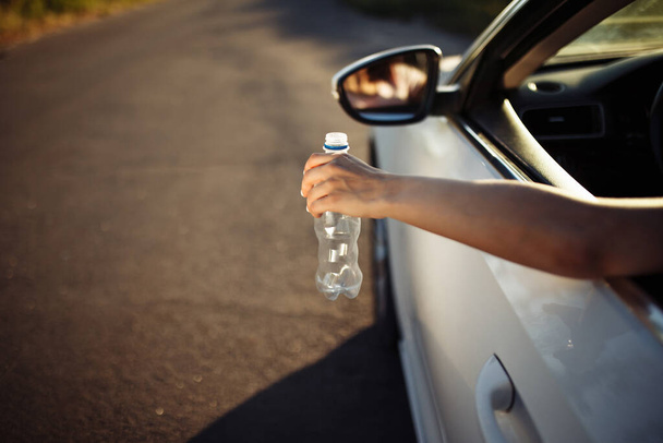 La main d'une femme jetant une bouteille en plastique par la fenêtre de la voiture. Le conducteur jette une bouteille d'eau usée sur la route. Pollution irresponsable de l'environnement et concept des ordures - Photo, image