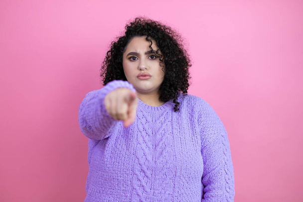 Молодая красивая женщина носит случайный свитер на изолированном розовом фоне указывая пальцем на камеру и для вас, уверенный жест выглядит серьезным - Фото, изображение