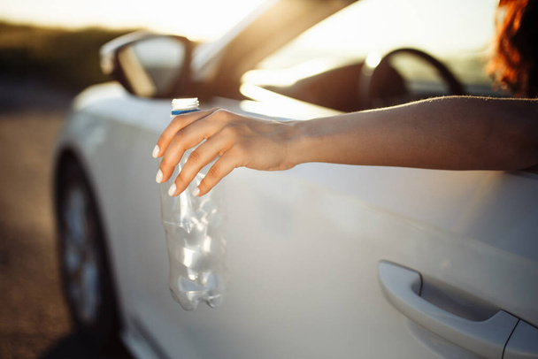 Ręka kobiety wyrzucającej plastikową butelkę przez okno samochodu. Kierowca wyrzuca zużytą butelkę wody na drogę. Nieodpowiedzialne zanieczyszczenie środowiska i koncepcja odpadów - Zdjęcie, obraz