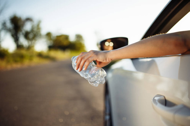 女性の手は車の窓の外にペットボトルを投げます。ドライバは、道路上で使用される水ボトルをスローします。環境やゴミの概念に対する責任のない汚染 - 写真・画像