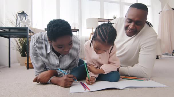 Afro-Amerikaanse ouders drie generaties grootvader, moeder en dochter kleindochter tekenen thuis op de vloer. volwassen moeder middelbare leeftijd vrouw volwassen afrikaanse man vader helpen klein meisje met huiswerk - Video