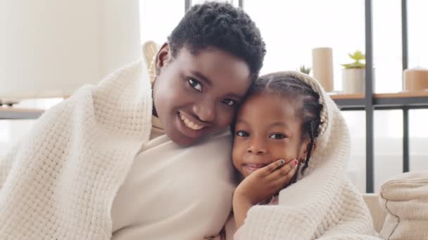 Portret afro amerykańskiej rodziny w domu pokryty kocem, afrykański samotny matka dorosły czarny kobieta przytula przytula się do mało słodkie mieszane rasa dziewczyna dziecko córka patrząc na aparat ze szczęśliwymi twarzami - Materiał filmowy, wideo