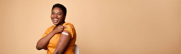 Mujer africana enrollándose las mangas mostrando brazo vacunado, fondo beige - Foto, imagen