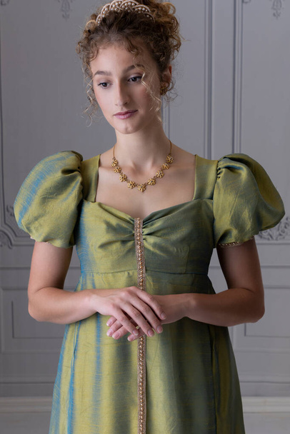 Una joven Regencia vistiendo un vestido de seda de tiro verde y de pie sola en una habitación con paredes de paneles  - Foto, Imagen