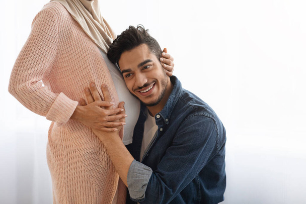 Homem oriental feliz abraçando a barriga de sua esposa grávida, ouvindo chutes de bebê - Foto, Imagem