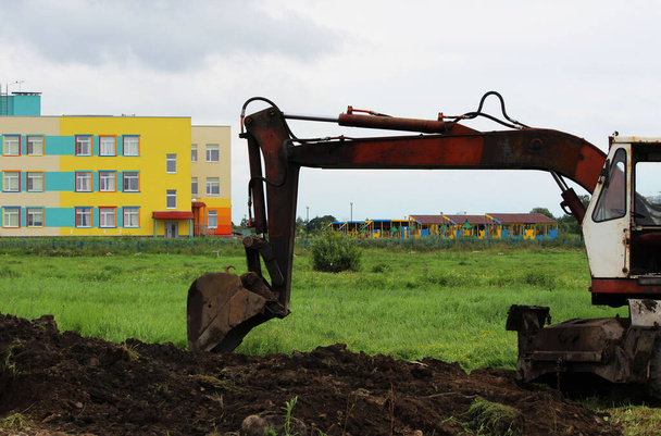 egy kotró ássa az árkot egy üres telken egy színes ház hátterében. Csőfektetés. riportkészítés. - Fotó, kép