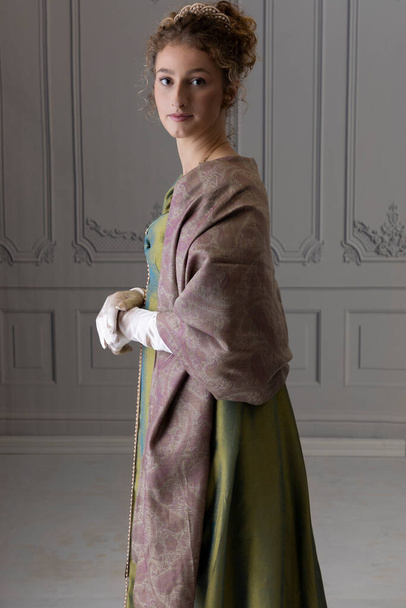 Una donna Regency indossa un abito di seta verde e uno scialle paisley in piedi da sola in una stanza con pareti rivestite di bianco - Foto, immagini