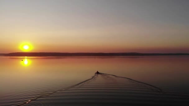 Disparo desde un dron de una puesta de sol de verano en un gran lago rodeado de bosques y rastros de una lancha a motor en el agua - Imágenes, Vídeo