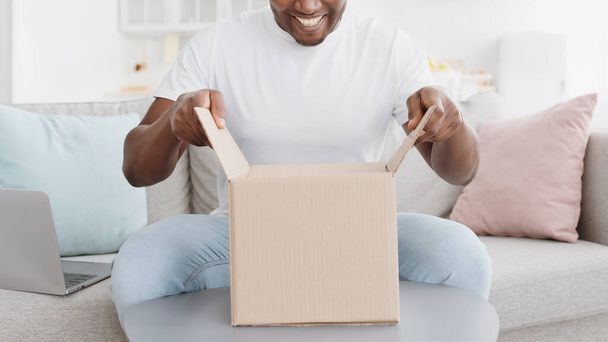 irreconocible hombre afroamericano desempacando caja de cartón, sentado en el sofá en casa - Foto, Imagen
