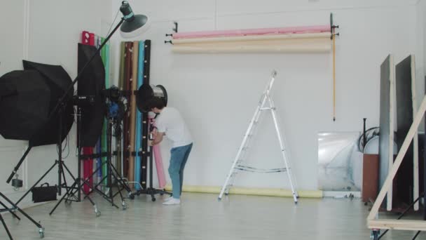 Ένας νεαρός άνδρας σε ένα στούντιο σχεδιασμού διαδίδει το φόντο για φωτογραφίες - ελαφρύ εξοπλισμό γύρω - Πλάνα, βίντεο
