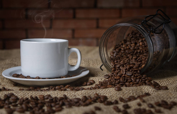 xícara de café e um frasco de vidro com grãos de café espalhados na mesa - Foto, Imagem