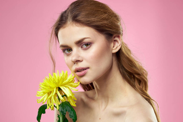 mooi meisje met een gele bloem op een roze achtergrond naakt schouders make-up - Foto, afbeelding