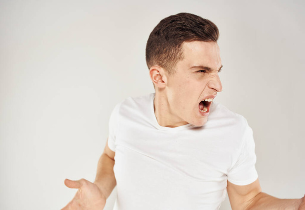 Un hombre con una camiseta blanca hace gestos con las manos sobre un fondo gris irritabilidad estrés  - Foto, Imagen