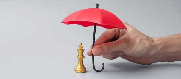 Деловой человек, держащий в руках красный зонтик, прикрывает фигурку Короля шахмат. Бизнес, управление рисками, решение, экономический регресс, страхование, стратегия и прерывание деятельности - Фото, изображение