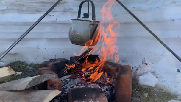 Télen egy turista teáskanna lóg egy égő tábortűz felett a természetben, ételt és teát főzve a tűzön. 4K - Felvétel, videó