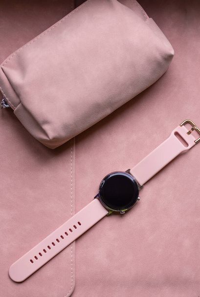 Современные гаджеты для девочек в мягком розовом цвете. Умные часы с розовым ремешком лежат на розовом чехле для ноутбука рядом с розовой косметической сумкой - Фото, изображение