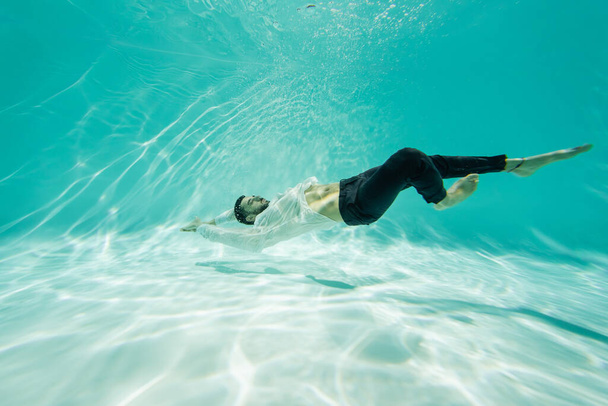 Hombre árabe en camisa blanca nadando bajo el agua en la piscina  - Foto, Imagen