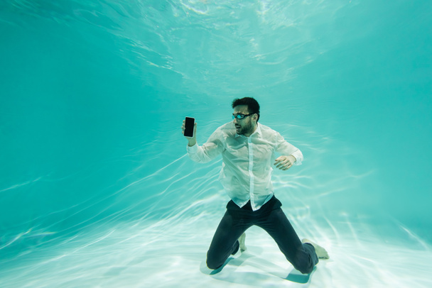 Arabian businessman in formal wear and goggles holding smartphone with blank screen underwater  - Photo, Image
