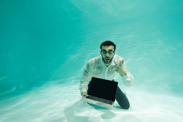 Eccitato uomo d'affari arabo che tiene il computer portatile con schermo vuoto sott'acqua  - Foto, immagini