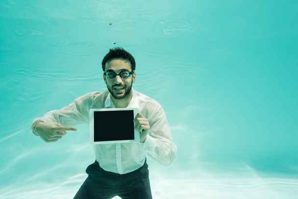 Hombre de negocios árabe sonriendo y apuntando a la tableta digital con pantalla en blanco bajo el agua  - Foto, Imagen