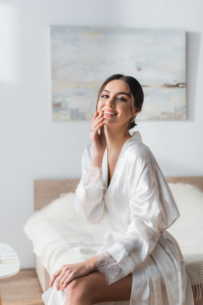 brunette bride in silk robe smiling at camera while touching face in bedroom - Foto, afbeelding