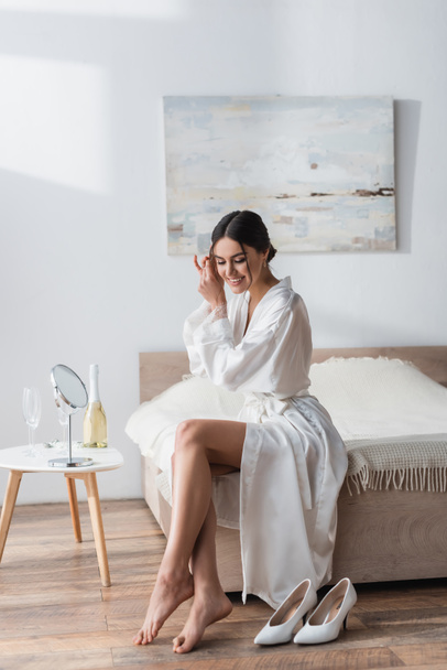 smiling bride in white robe fixing hair near mirror and champagne on bedside table - Foto, afbeelding
