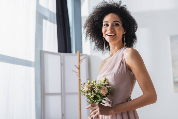 cheerful african american bridesmaid looking away while holding wedding bouquet - Fotó, kép