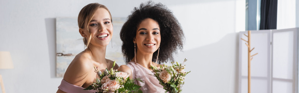結婚式の花束、バナーを保持しながらカメラを見て幸せな異人種間の新婦付添人 - 写真・画像