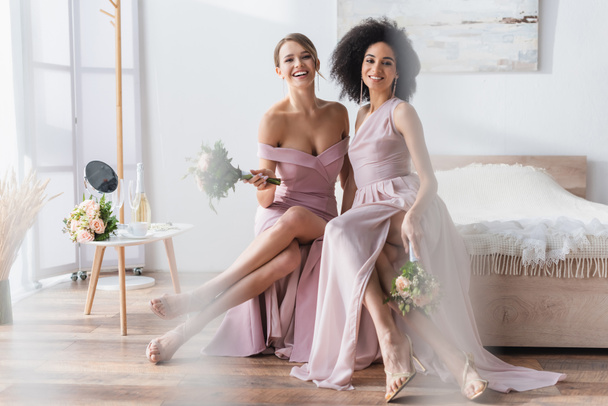 charming multicultural bridesmaids sitting on bed with wedding bouquets - Photo, image