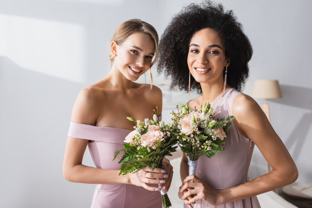 pretty multiethnic bridesmaids holding wedding bouquets and smiling at camera - Fotó, kép