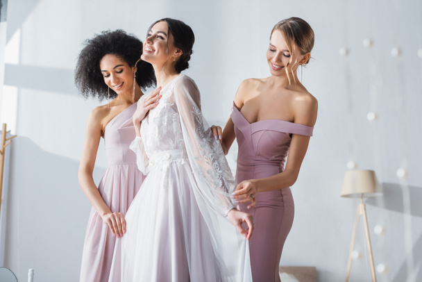 joyful bride holding hand on chest while multicultural bridesmaids adjusting her dress - Fotografie, Obrázek