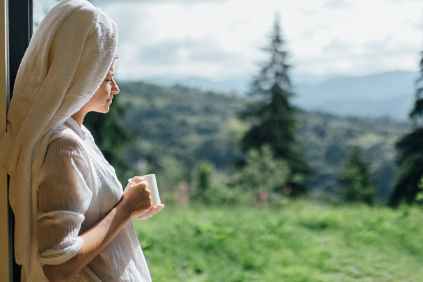 Hermosa mujer joven disfrutando del café en la mañana soleada en el fondo de las colinas de montaña. Tranquilo momento tranquilo. Hermosa hembra en pijama blanco y toalla después de la ducha relajante con taza de café - Foto, Imagen