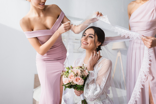 bridesmaids holding veil over pleased bride with wedding bouquet in bedroom - Foto, Imagen
